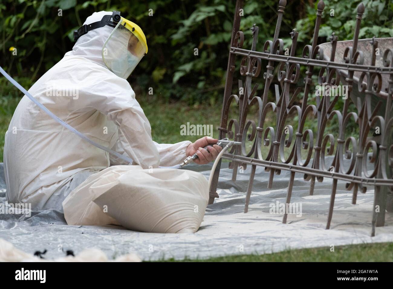 Sabbiatura di una vecchia recinzione metallica. Un uomo in una tuta bianca e una maschera antipolvere. Pulizia e restauro di elementi metallici, Foto Stock