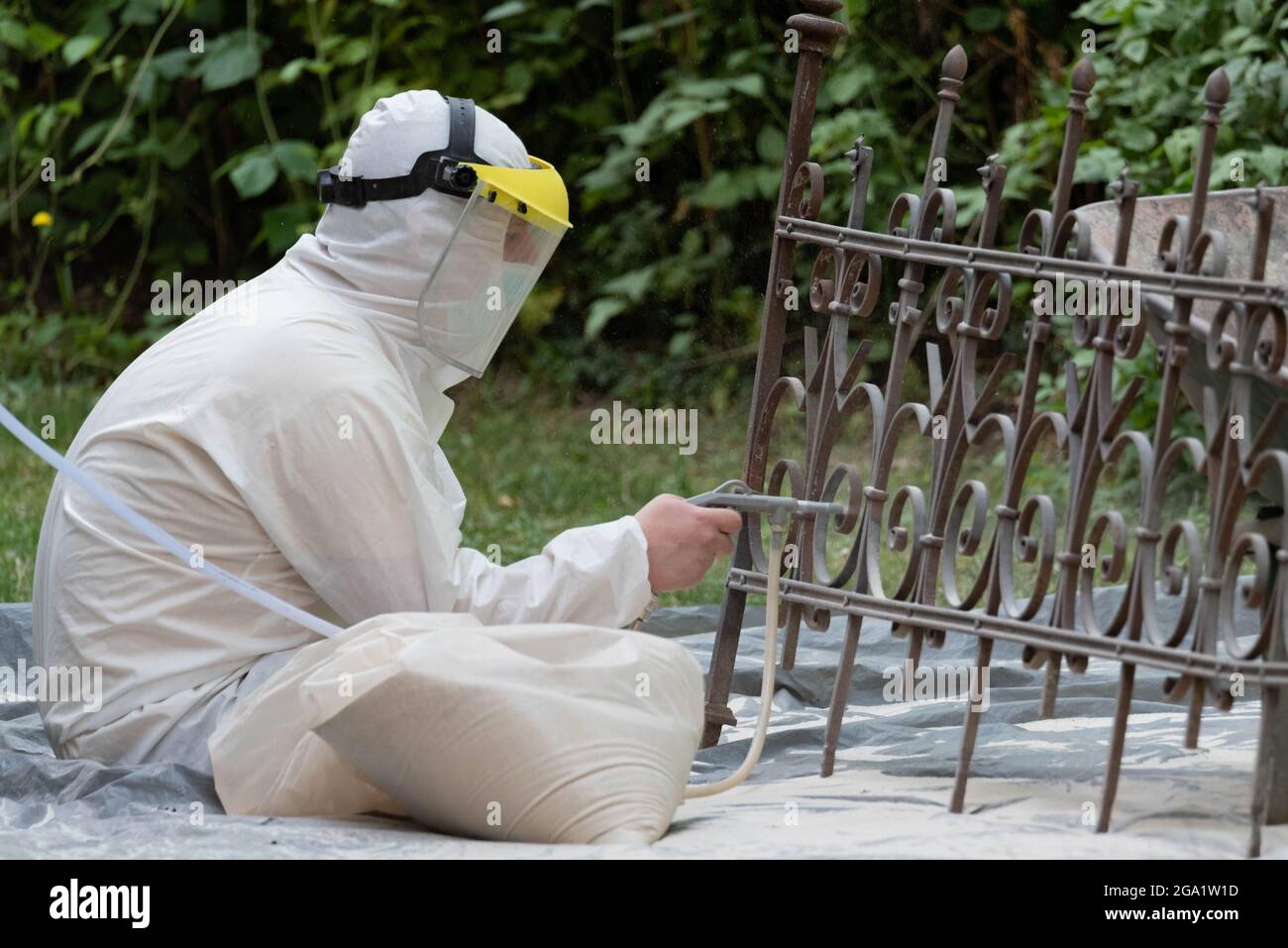 Sabbiatura di una vecchia recinzione metallica. Un uomo in una tuta bianca e una maschera antipolvere. Pulizia e restauro di elementi metallici, Foto Stock