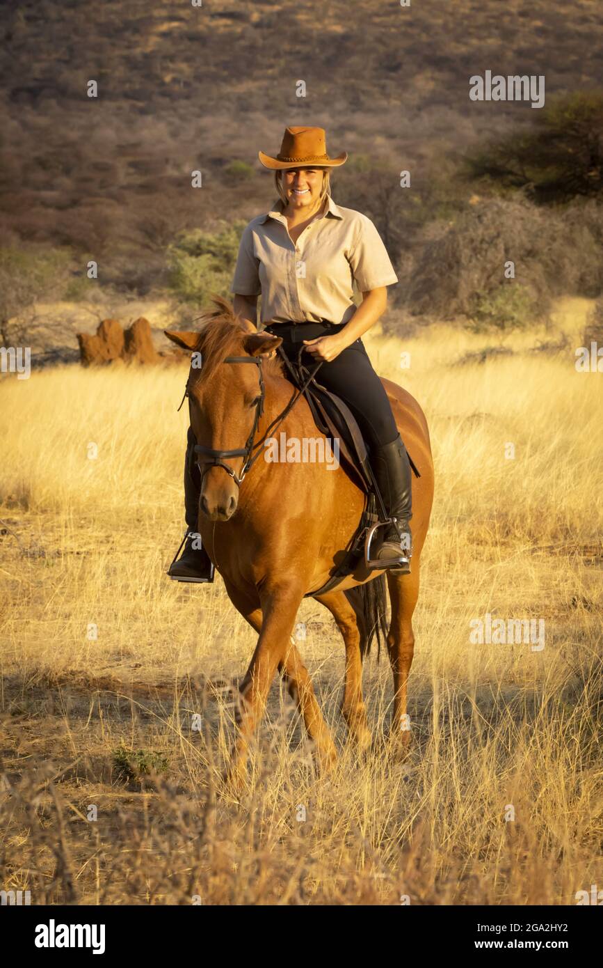 Donna a cavallo (Equus ferus caballus) attraverso il cespuglio sulla savana con la calda luce del tramonto, guardando la macchina fotografica e sorridendo il G... Foto Stock