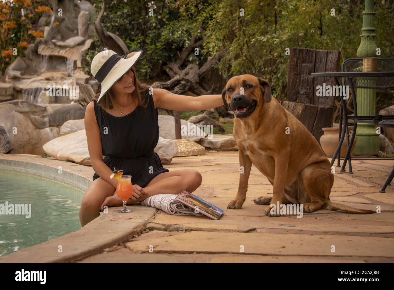 Donna che indossa un cappello da sole seduto in piscina e accarezza un cane (Canis lupus familiaris) al Gabus Game Ranch; Otavi, Otjozondjupa, Namibia Foto Stock
