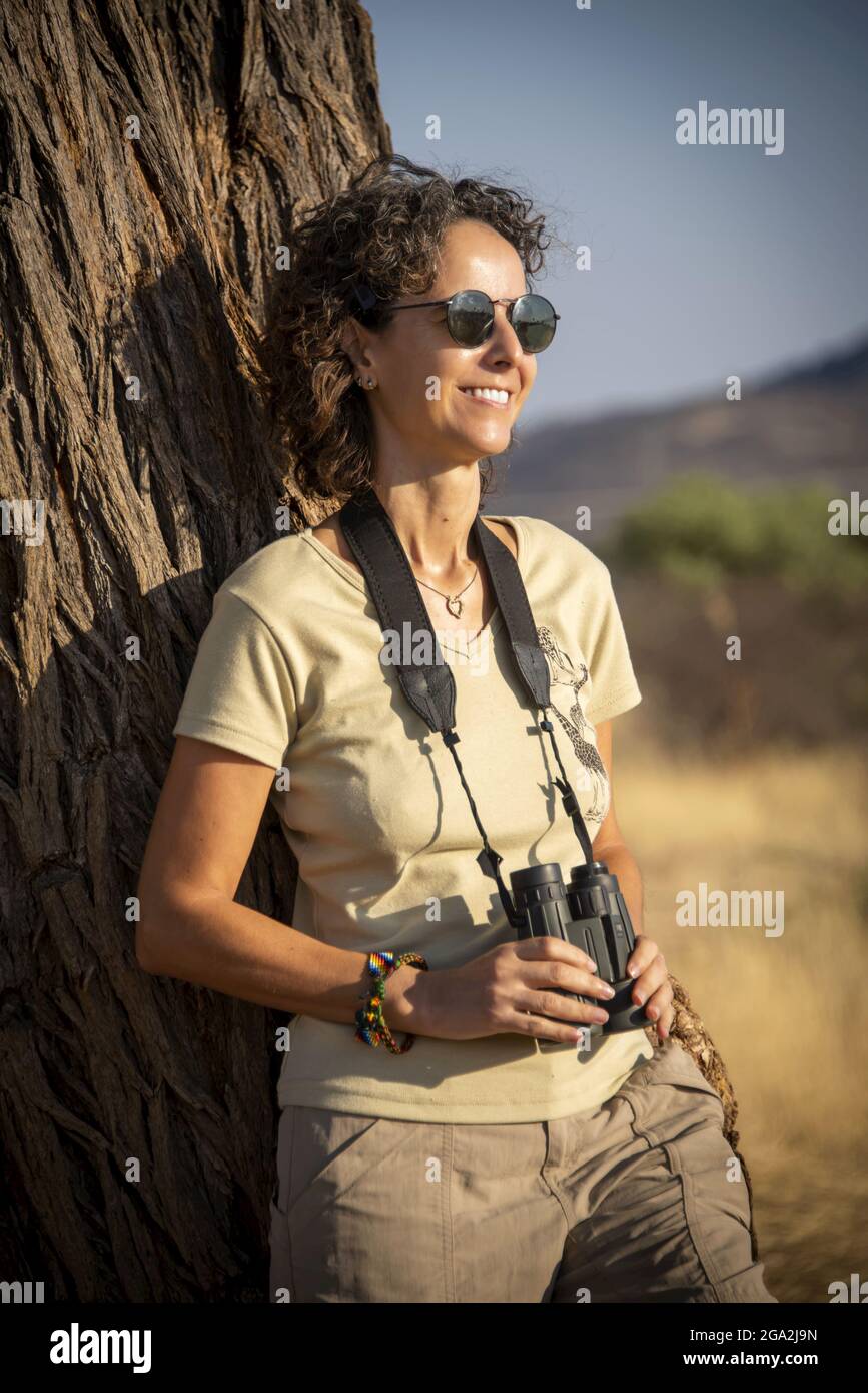Ritratto di una donna appoggiata su un tronco di albero indossando pantaloncini e occhiali da sole e tenendo un paio di binocoli mentre si guarda... Foto Stock