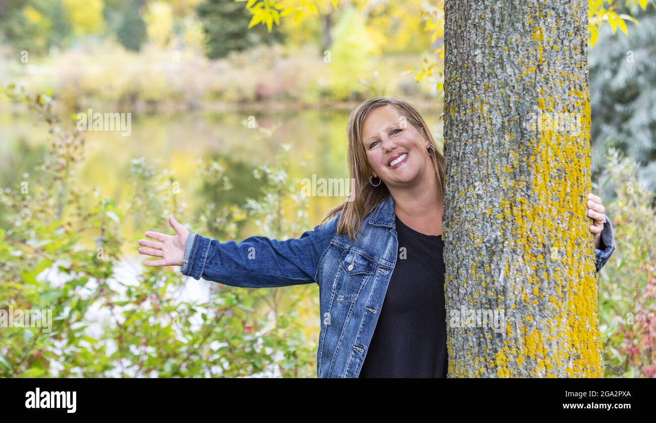 Una donna di mezza età che tiene un albero e raggiunge la sua mano mentre si gode in una calda serata autunnale in un parco cittadino; Edmonton, Alberta, Canada Foto Stock