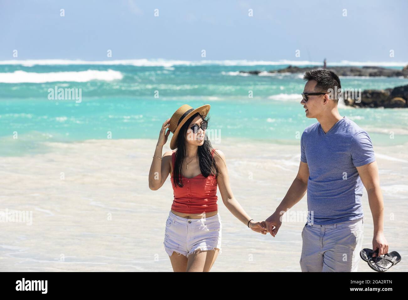 Una coppia che cammina su una spiaggia di sabbia bianca tenendo le mani e parlando insieme mentre si gode una vacanza tropicale lungo la costa di Oahu, Hawaii Foto Stock