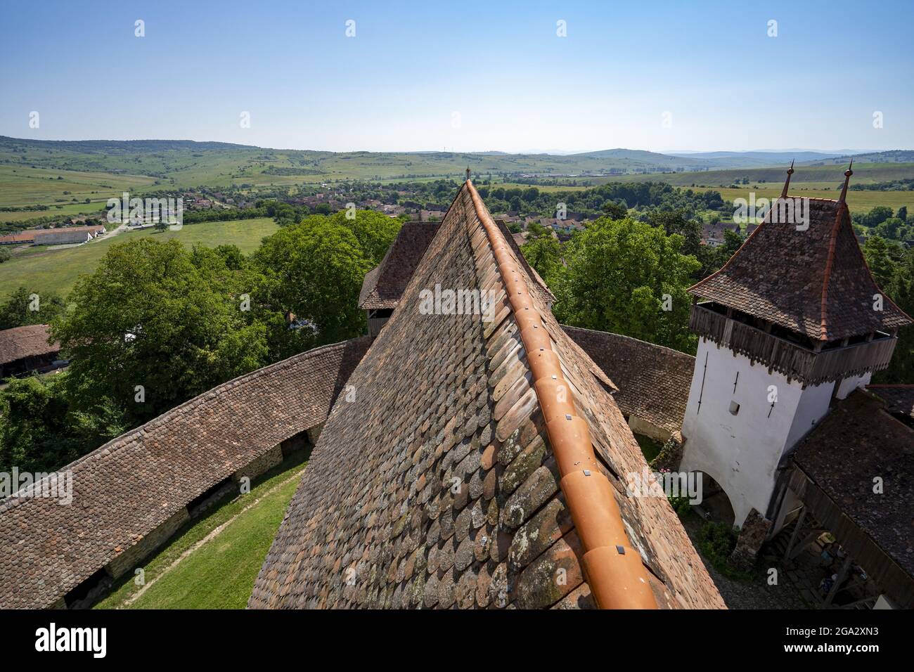 Primo piano del tetto piastrellato della Chiesa sassone fortificata di Viscri con le sue mura in pietra circostanti e una panoramica del paese e della campagna Foto Stock