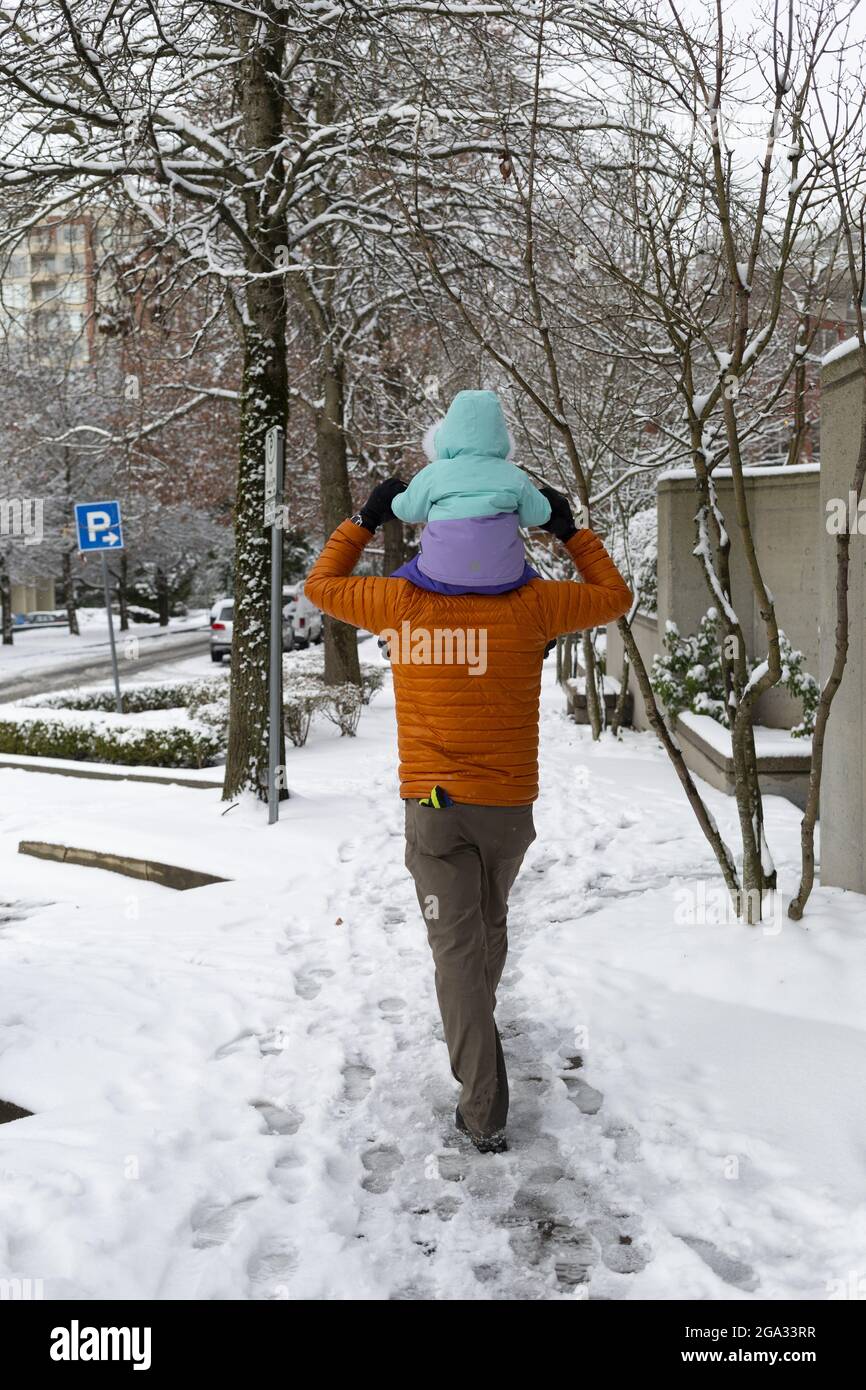 Padre cammina lungo un marciapiede dando un giro in spalla a sua figlia il giorno nevoso; North Vancouver, British Columbia, Canada Foto Stock