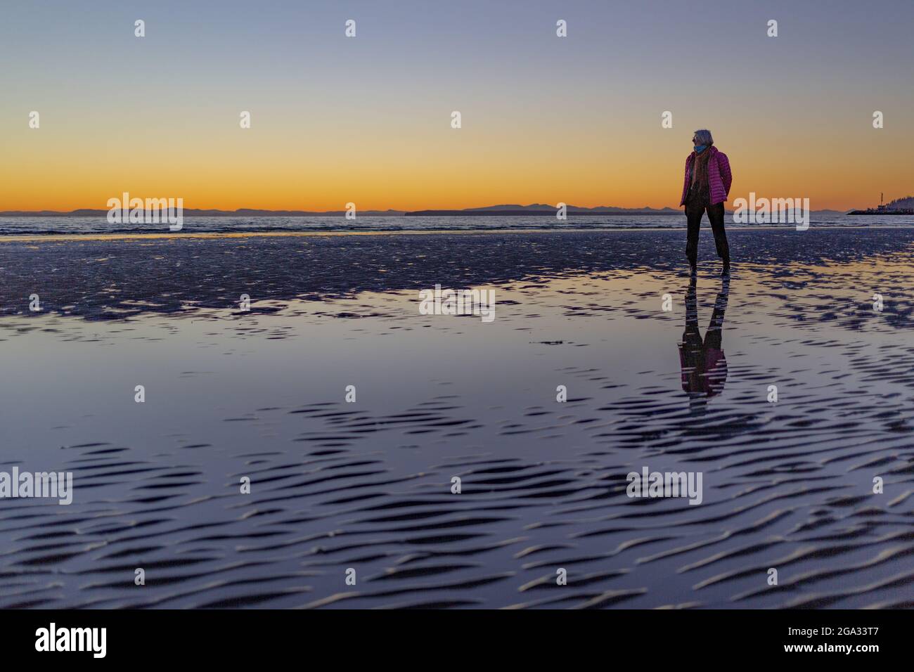 Una donna anziana si erge su una spiaggia bagnata che guarda fuori al cielo del tramonto e dell'oceano con una maschera per il viso tirata giù dal suo viso, raffigurante... Foto Stock