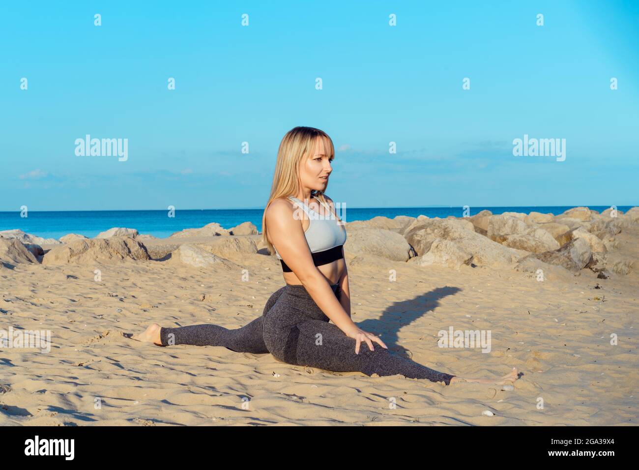 Giovane donna fitness in abbigliamento sportivo facendo stretching seduta su spago nella spiaggia di sabbia durante l'allenamento all'aperto. Stile di vita attivo e assistenza sanitaria. Copia Foto Stock