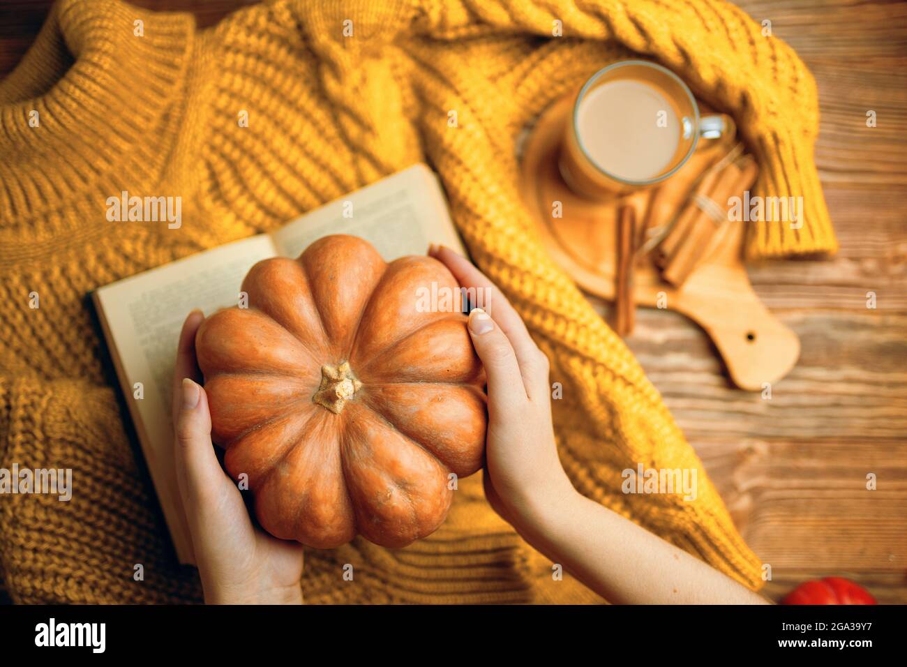 Zucca matura nelle mani delle donne sopra il maglione lavorato a maglia. Cioccolata calda con bastoncini di cannella su vassoio di legno. Pullover arancione, libro romantico aperto e zucca su sfondo di legno. Composizione autunnale. Foto Stock