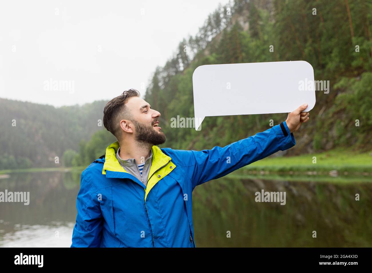 Il giovane uomo con barba in giacca dice qualsiasi parola con possibilità di scriverle su sfondo bianco rettangolare, che tiene con la mano contro b Foto Stock