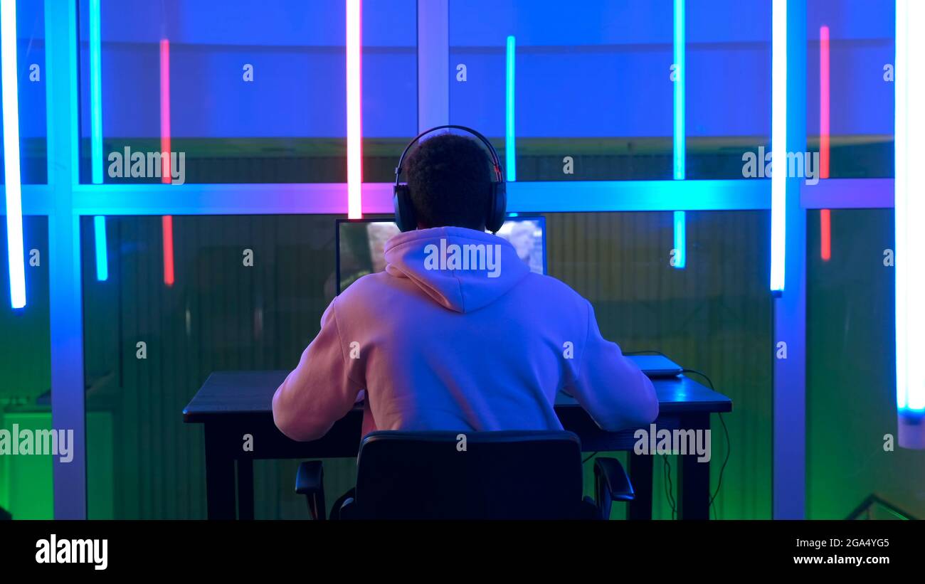 Videogamer professionista di razza mista che gioca nel videogioco online sparatutto su pc in una grande stanza con luci LED dai colori caldi. Foto Stock