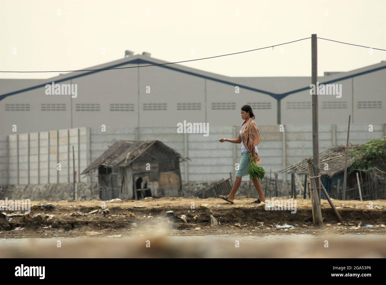 Una donna cammina sul lato del canale delle inondazioni di Giacarta, in uno sfondo di edifici di magazzino in una nuova area commerciale chiamata Marunda Makmur che amministrativamente si trova nella reggenza di Bekasi, Giava Occidentale, Indonesia. Fotografato nel villaggio di Marunda, Cilincing, Giacarta Nord, Giacarta, Indonesia. Foto Stock