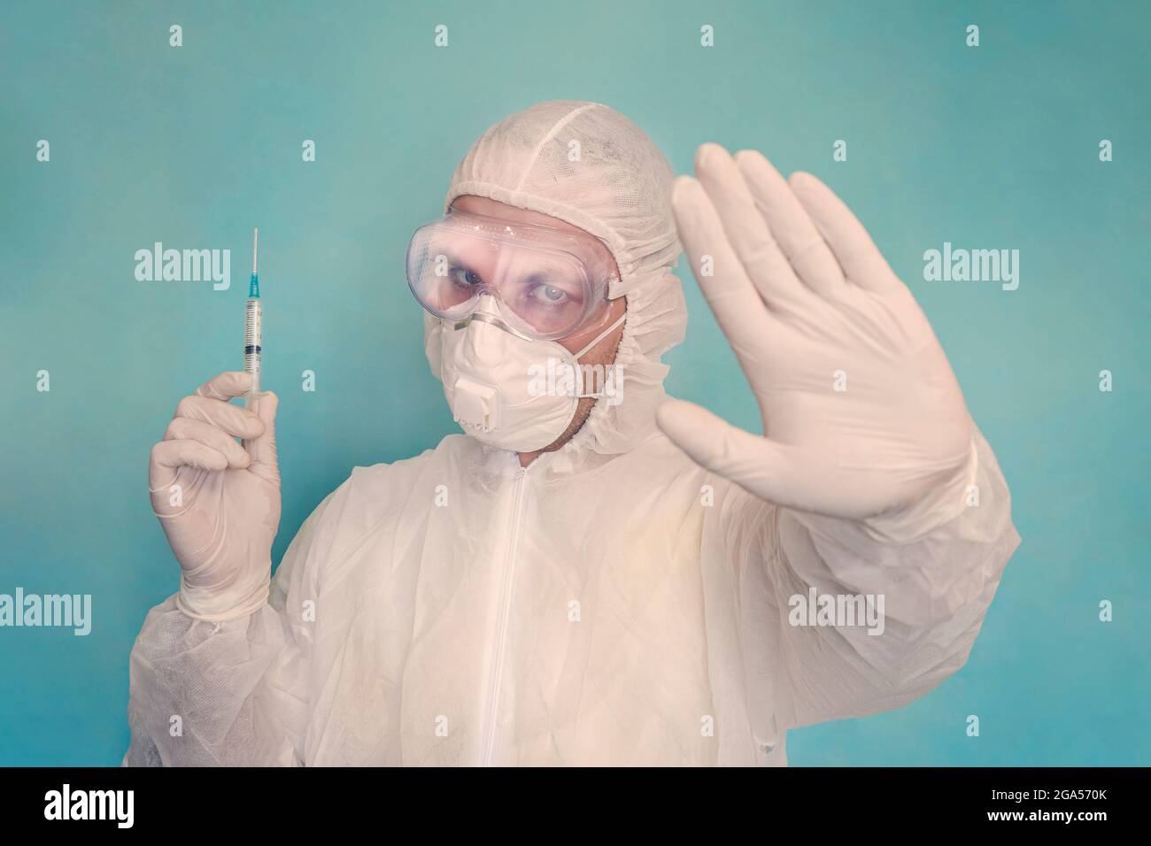 Ritratto del medico maschile in indumenti protettivi, guanti e maschera che tengono una siringa e mostrando un gesto proibitivo con le mani. Medicina e assistenza sanitaria co Foto Stock