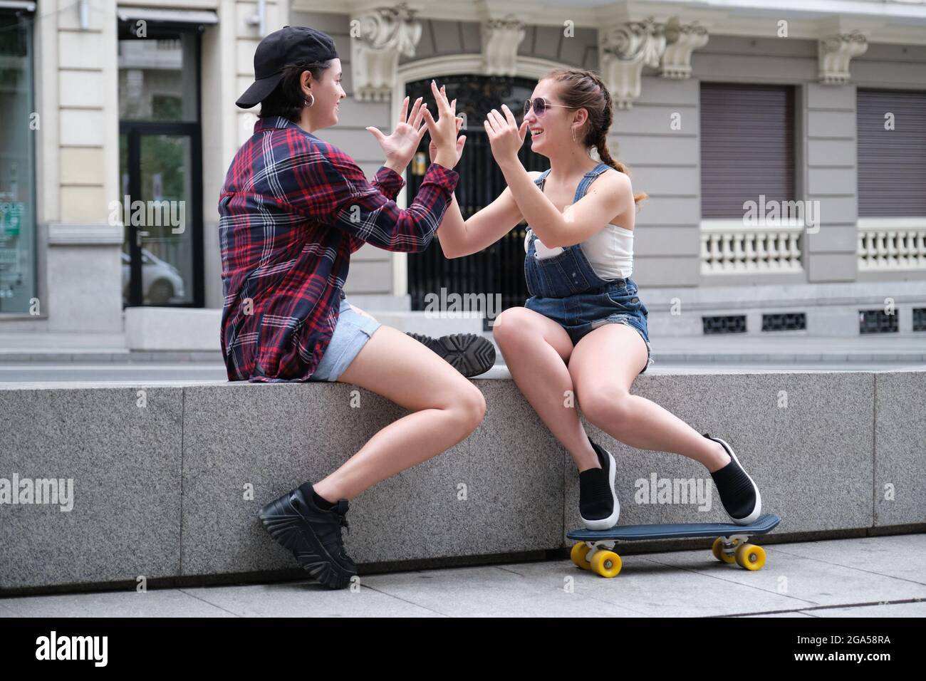 Due belle donne sorridenti, giocando una partita a mano, seduti in strada con uno skateboard. Foto Stock