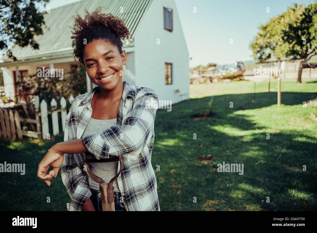 Razza mista agricoltore femminile in piedi in villaggio agricolo appoggiato su pala sorridente dopo una lunga giornata di lavoro Foto Stock