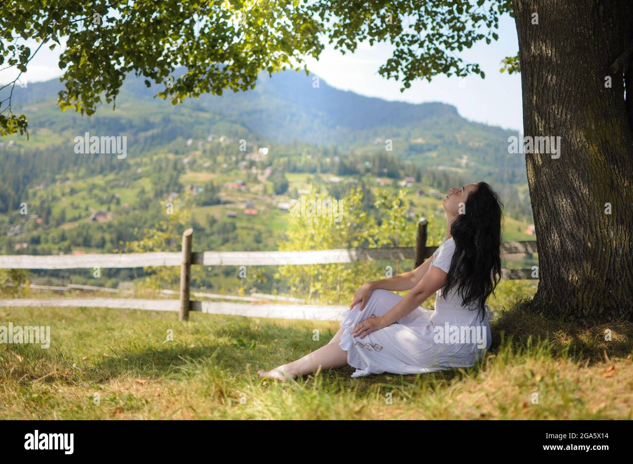 Giovane donna in abito bianco è seduta sotto un albero sul prato di montagna. Foto Stock