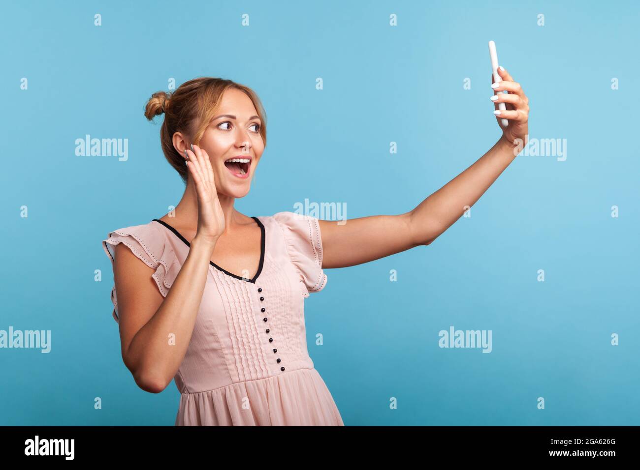 Vista laterale della donna felice amichevole con i capelli buni che prendono selfie e la mano ondeggiante, gesturing ciao per i seguaci, comunicando in videochiamata. Stu. Interno Foto Stock