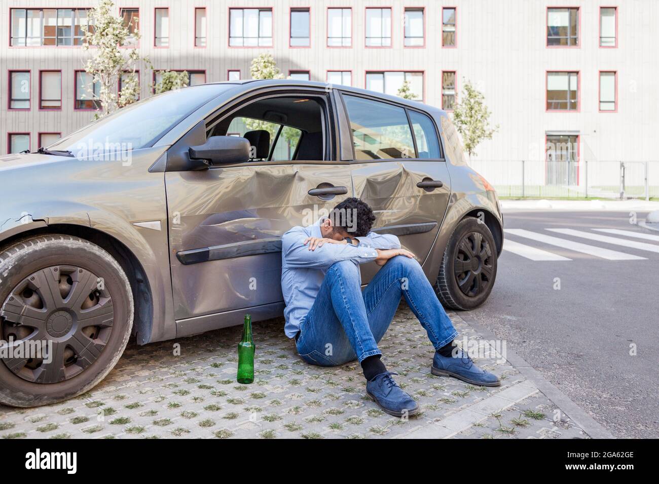 Uomo che indossa jeans e camicia blu seduto vicino auto schiantato con una bottiglia di bevande alcoliche e dormire, auto danneggiato ubriaco guida, violazione le Foto Stock