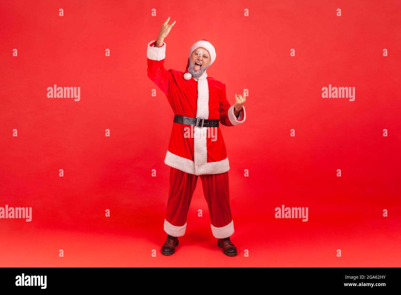 Foto a tutta lunghezza di un anziano entusiasta con barba grigia con costume di babbo natale che mostra rock e roll cantare con la macchina fotografica e urlare con un look pazzo Foto Stock