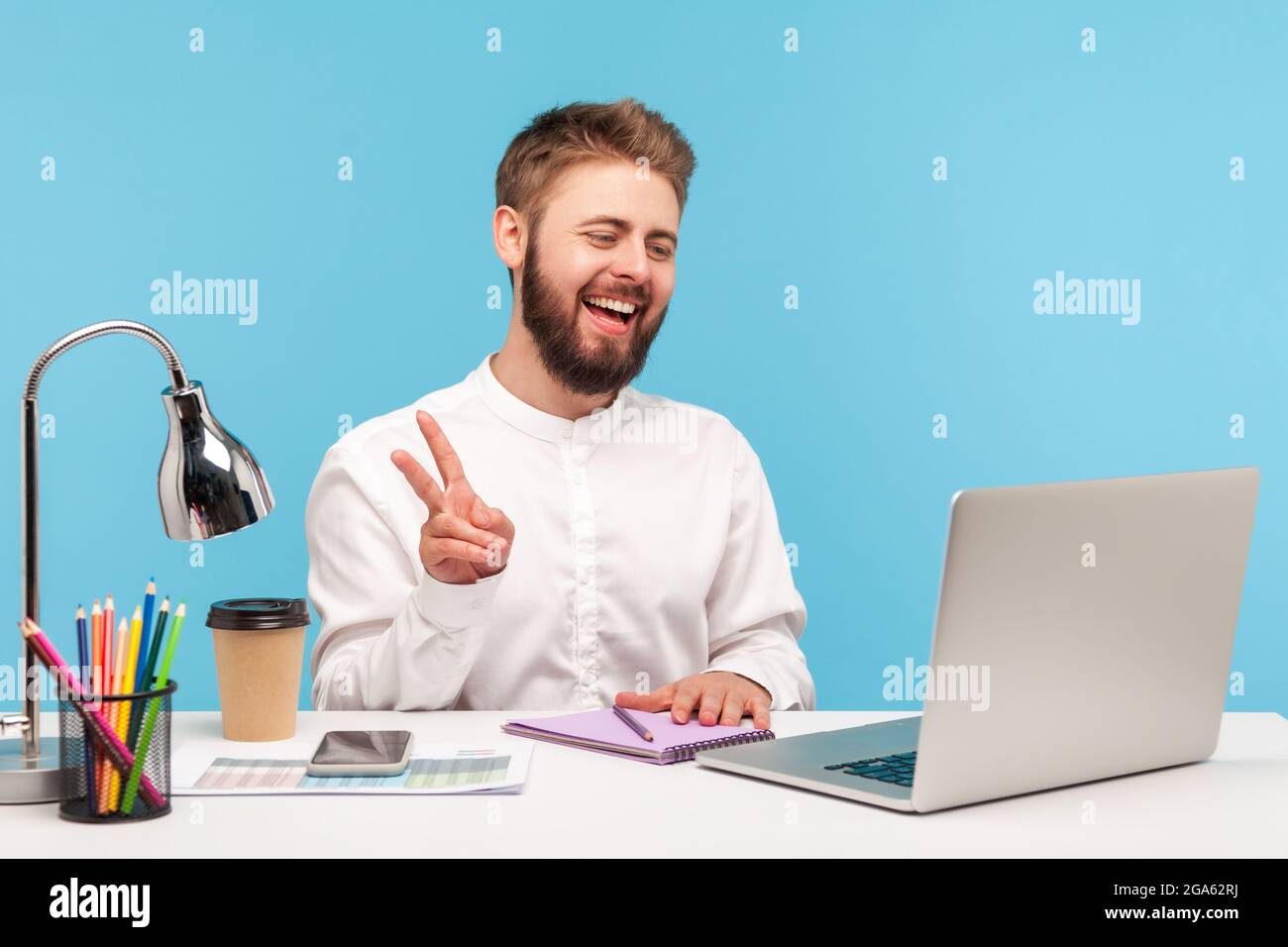 Un lavoratore di ufficio con la bearded positivo che mostra v gesture con le dita che parlano videochiamata, guardando la fotocamera del laptop con il sorriso toothy seduto sul posto di lavoro. Foto Stock