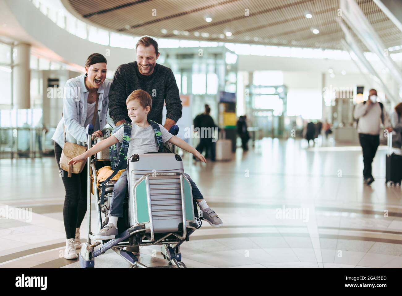 Coppia felice di spingere il trolley con il loro figlio all'aeroporto. I bambini si divertono seduti sul carrello dei bagagli mentre i genitori lo spingono all'aeroporto. Foto Stock