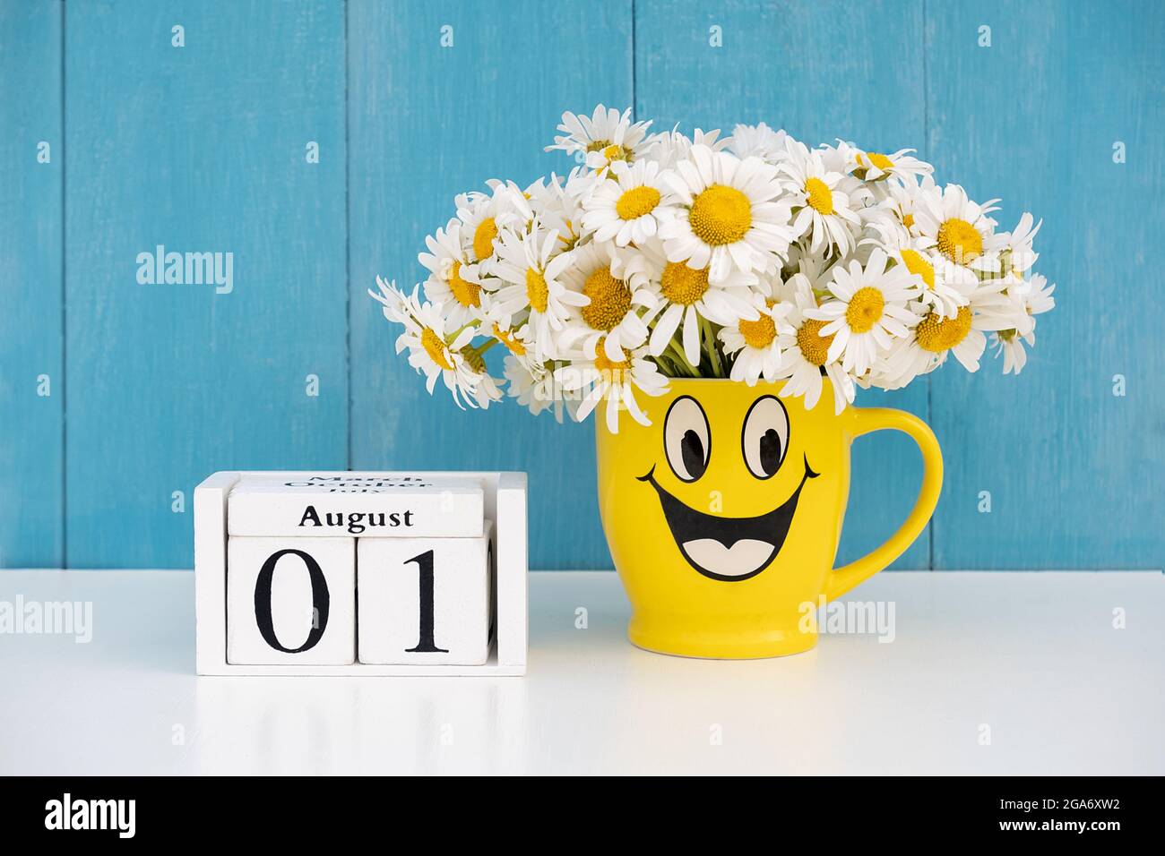 1 agosto data su calendario e bouquet di camomilla in tazza gialla con faccia divertente contro parete blu. Ciao agosto estate mese concetto. Foto Stock
