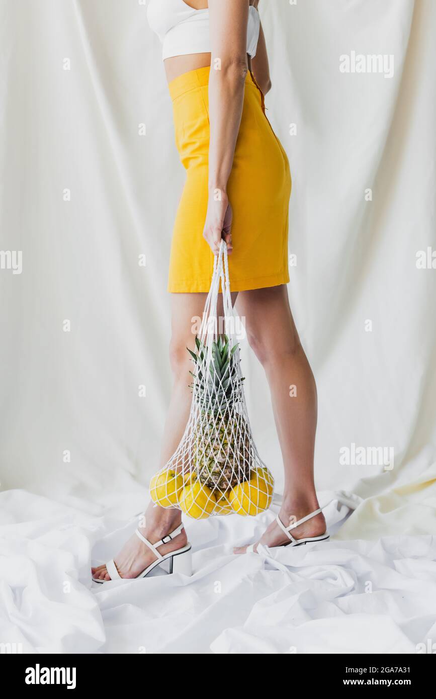 vista parziale della donna con gonna gialla e borsetta con filo di contenimento con limoni e ananas su bianco Foto Stock