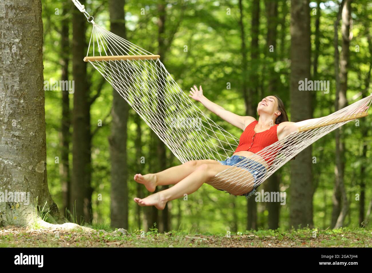 Donna eccitata che oscilla sull'amaca festeggiando la vacanza in una bella foresta verde Foto Stock