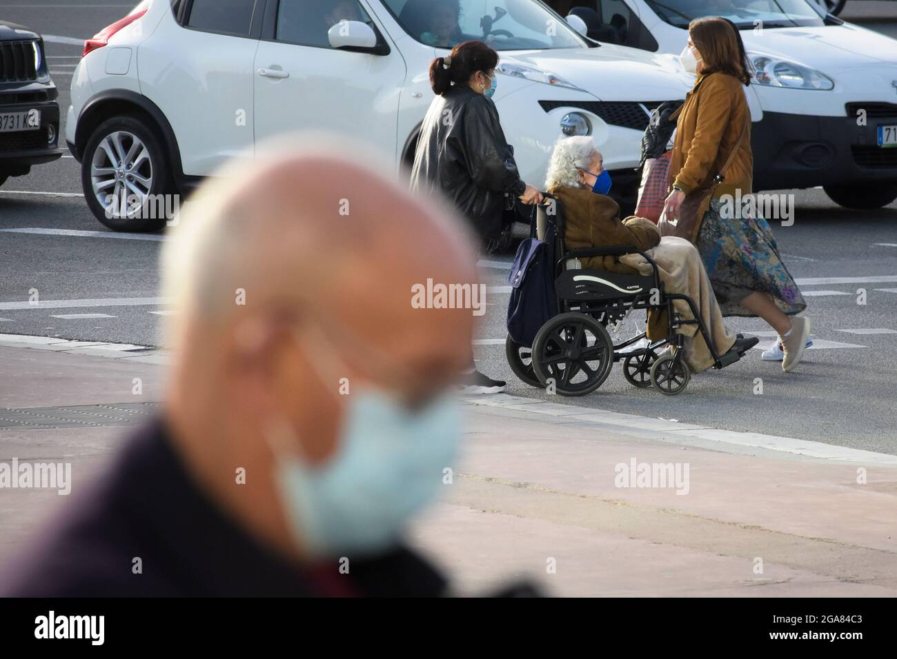 Persone che camminano in una strada con maschera durante i periodi di pandemia. L'operatore sanitario spinge la sedia a rotelle con una donna anziana seduta in essa. Foto Stock