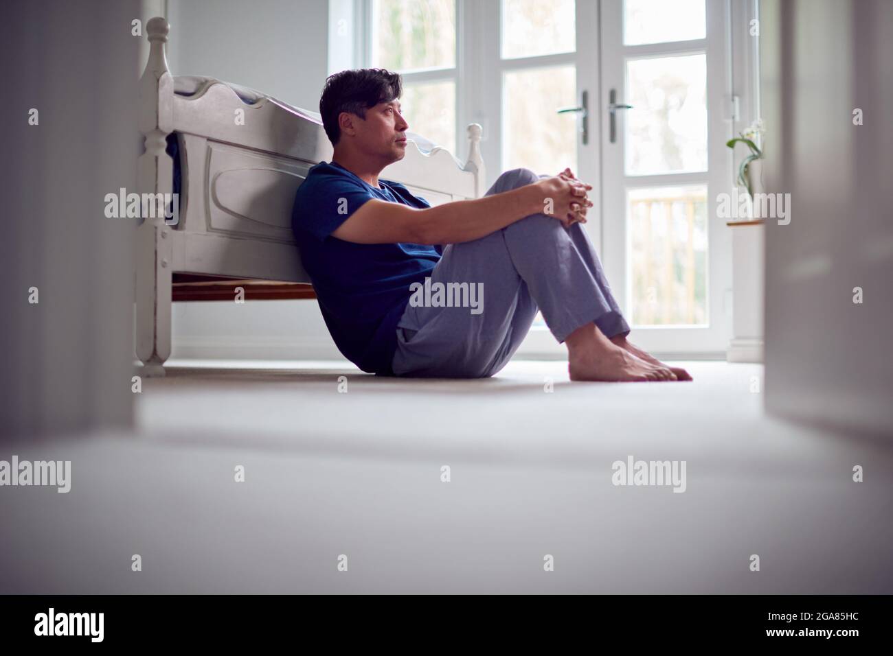 Infelice e depresso uomo asiatico maturo in Pajamas seduta sul pavimento della camera da letto Foto Stock