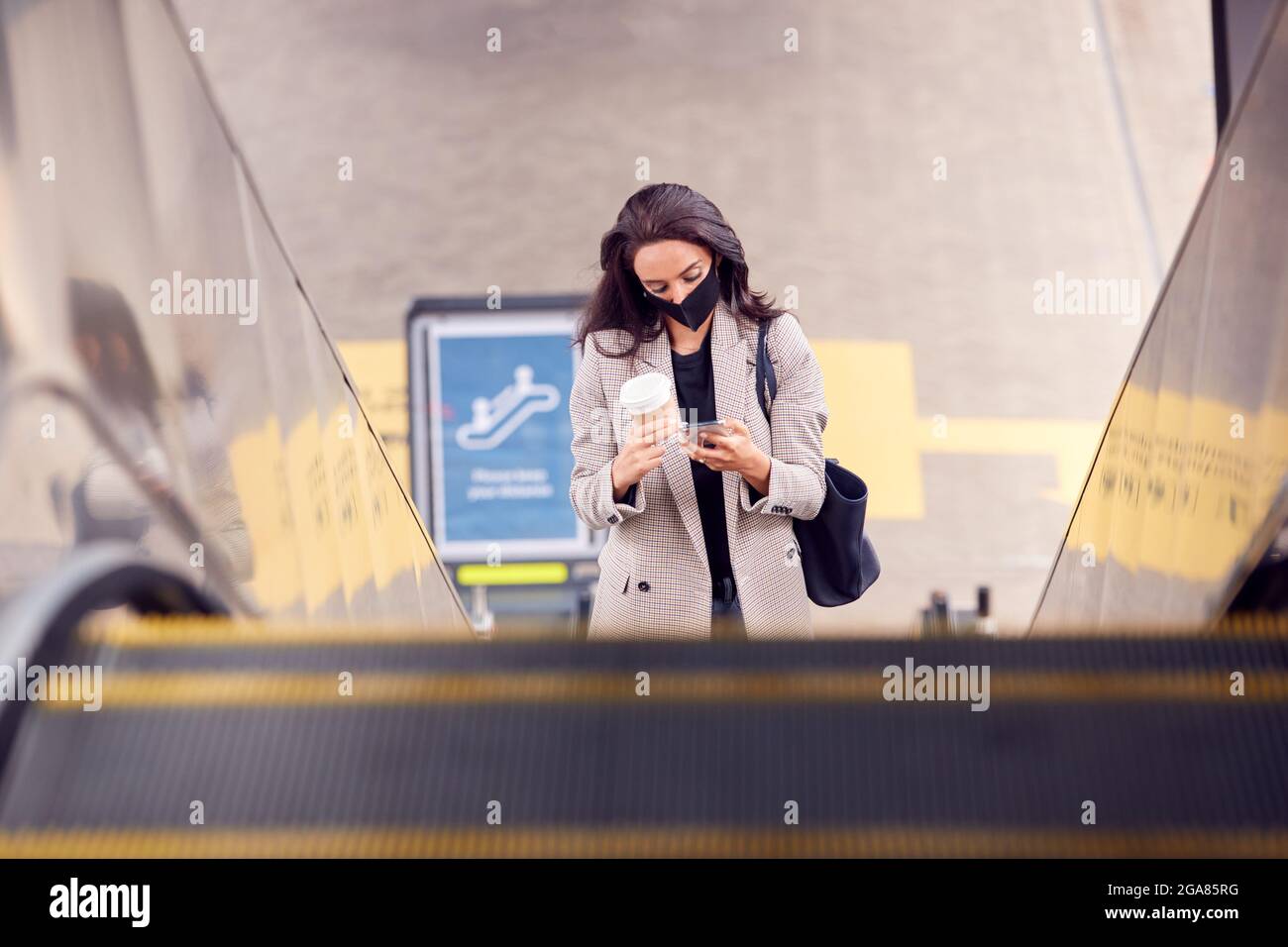 Donna d'affari su Escalator alla stazione ferroviaria con il telefono cellulare indossando la maschera DPI in Pandemic Foto Stock