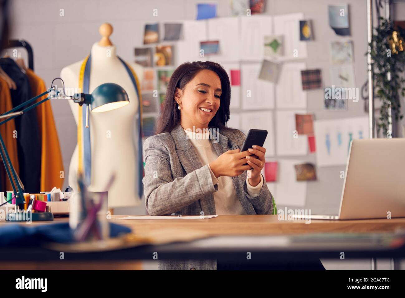Immagine di messa a fuoco differenziale del progettista di moda femminile usando il telefono mobile per navigare in Internet alla scrivania Foto Stock