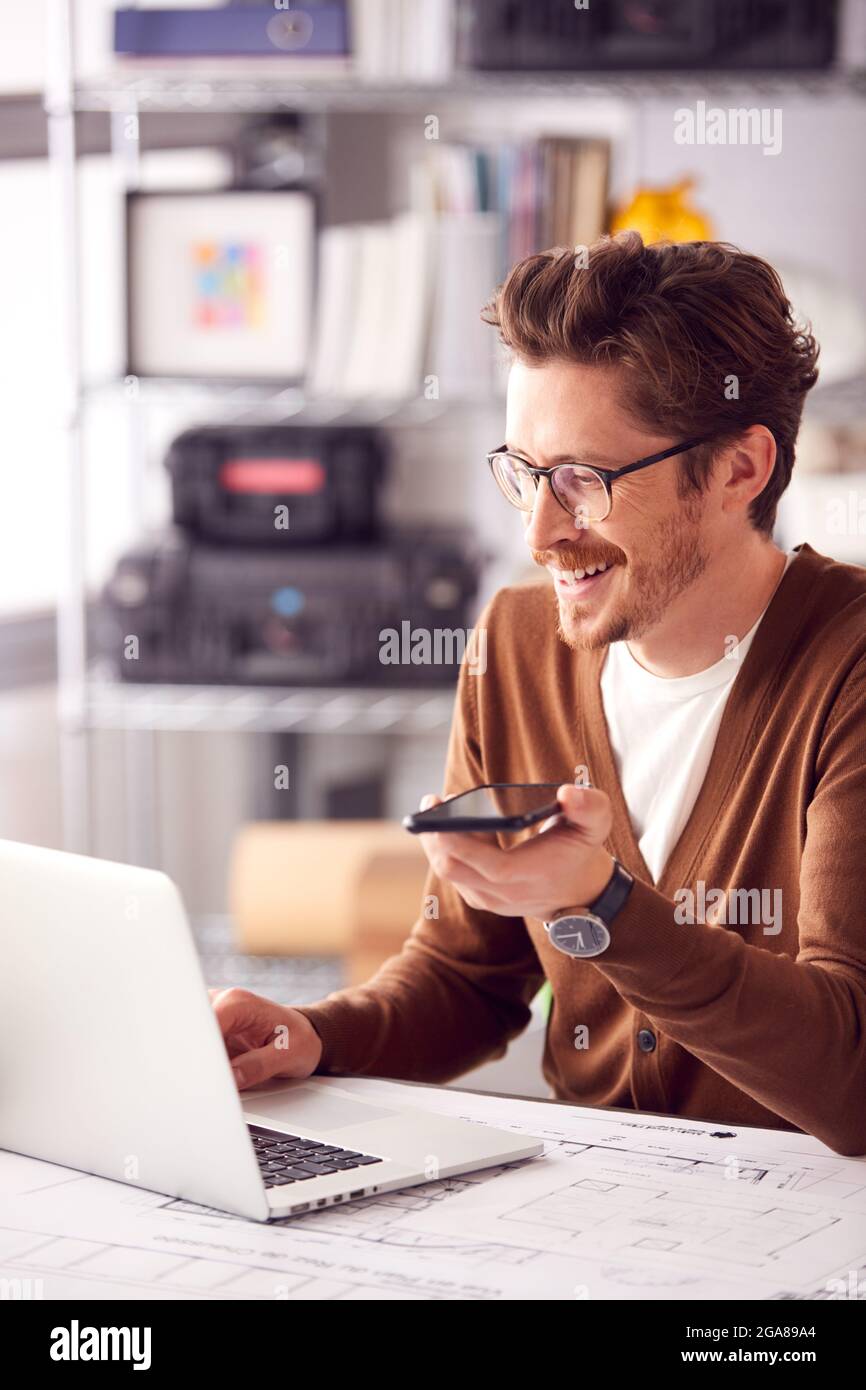 Architetto maschile in ufficio che lavora alla scrivania su laptop che prende la chiamata vivavoce sul telefono cellulare Foto Stock