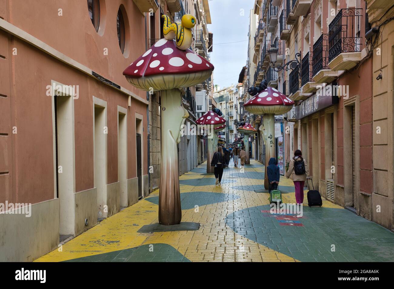Un vicolo lastricato con alti funghi giganti d'arte distanziato e la gente che cammina nella città di Alicante, Spagna Foto Stock