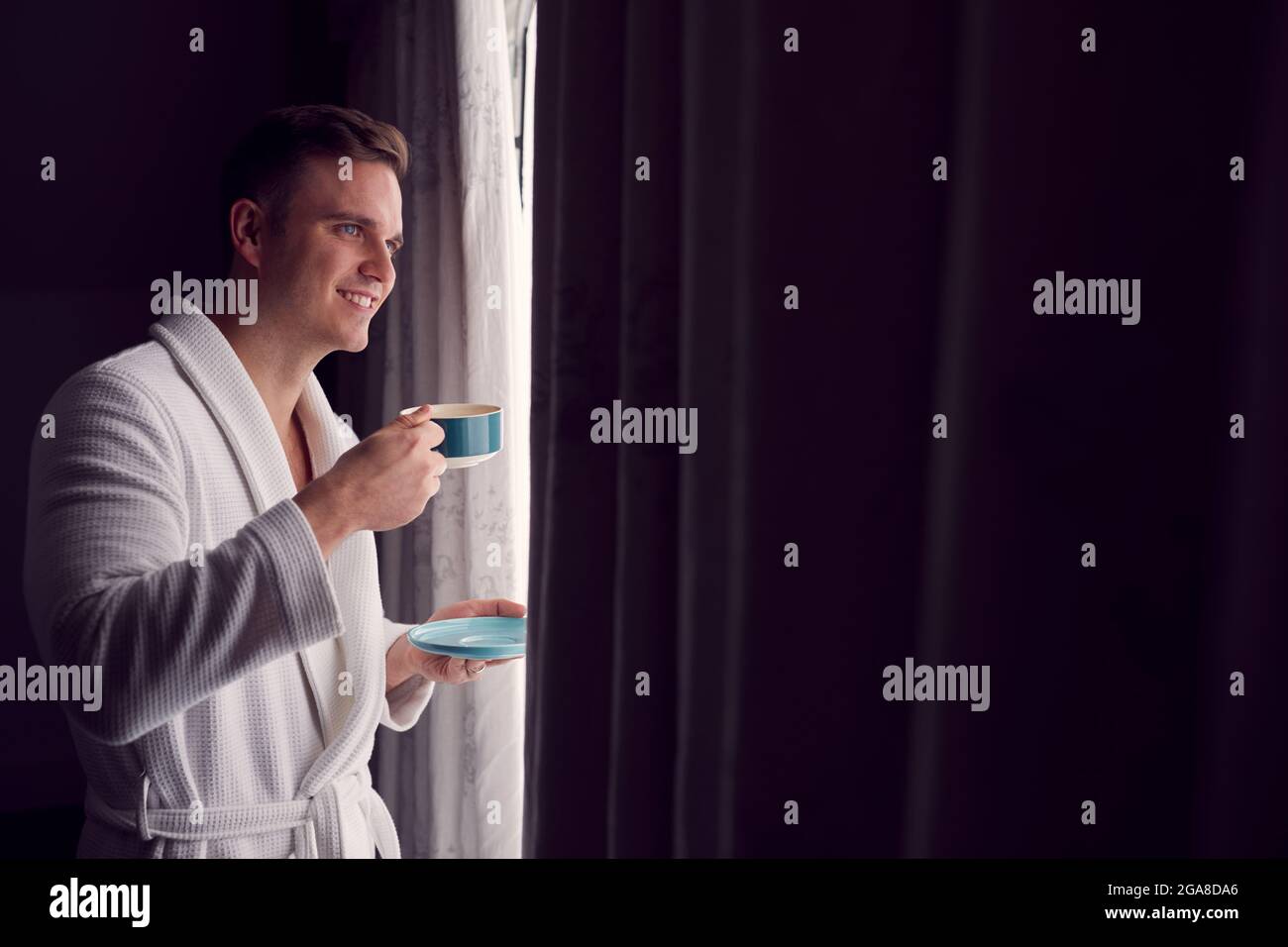 Uomo che indossa accappatoio in hotel o Spa Break Standing by Open Tende con bevanda calda Foto Stock