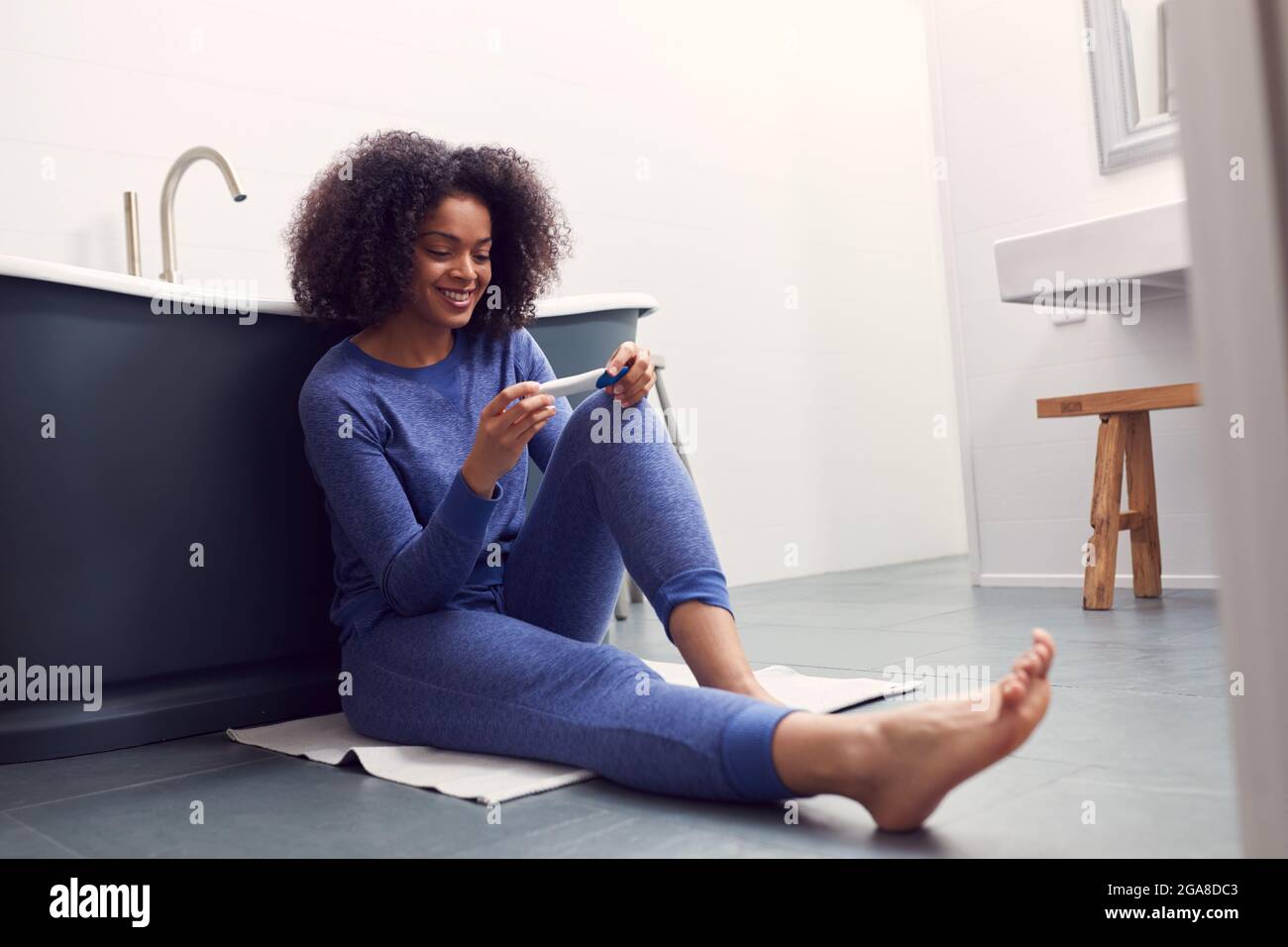 Donna eccitata seduta sul pavimento in bagno a casa con prova di gravidanza a casa positiva Foto Stock