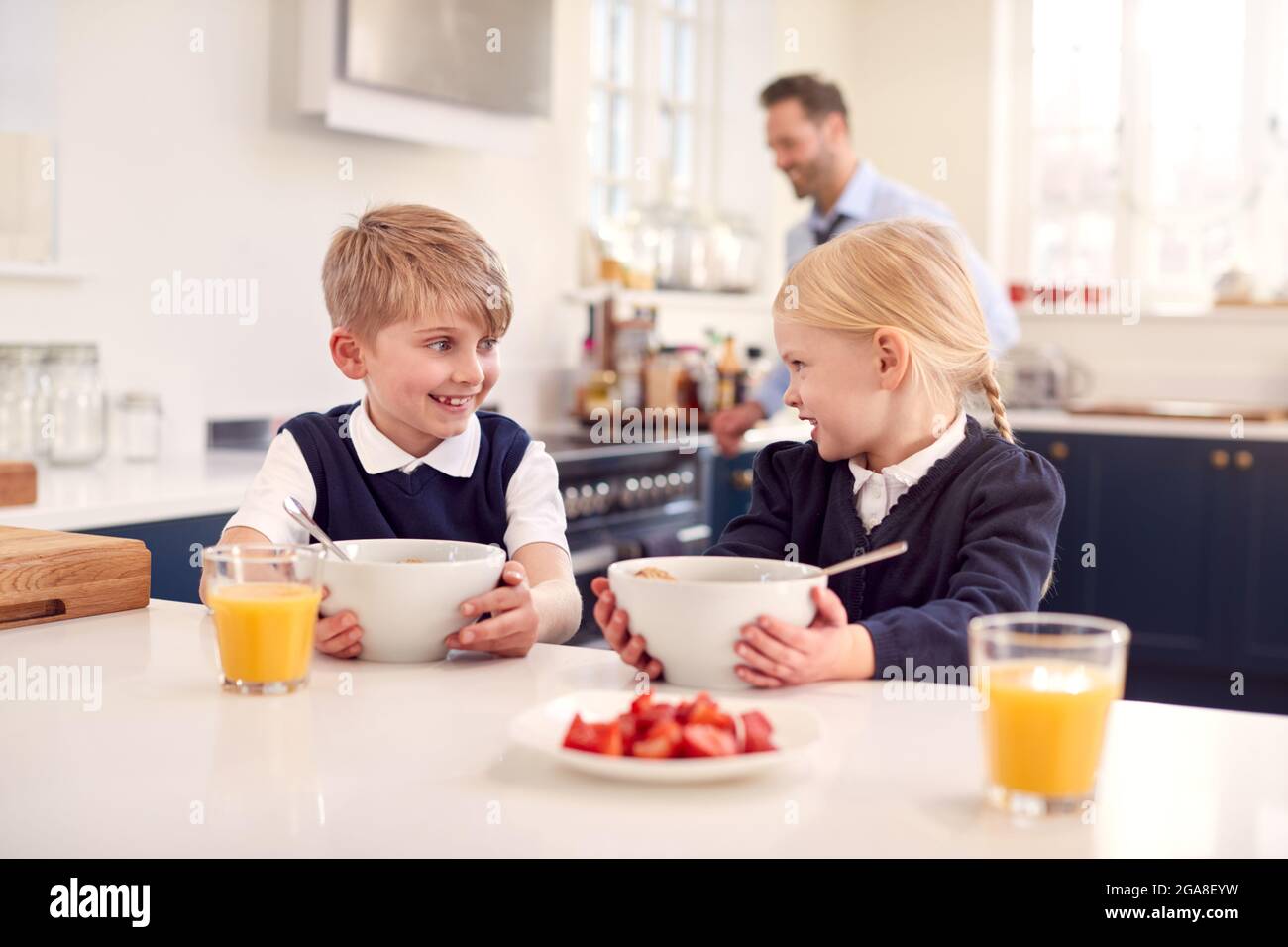 Due bambini che indossano l'uniforme della scuola in cucina che mangiano la colazione mentre il Padre si prepara per il lavoro Foto Stock