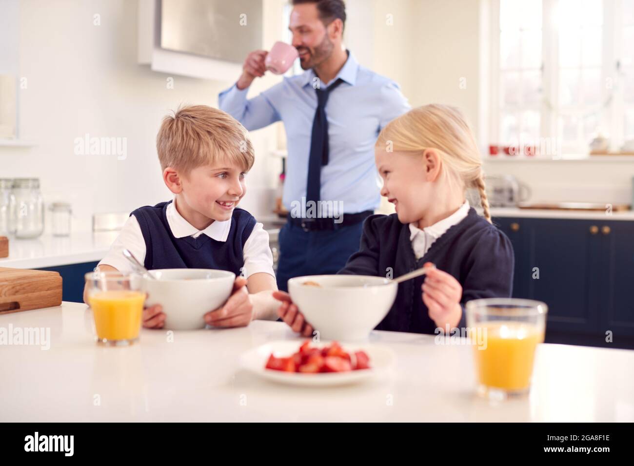 Due bambini che indossano l'uniforme della scuola in cucina che mangiano la colazione mentre il Padre si prepara per il lavoro Foto Stock