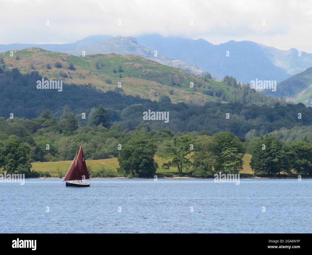Paesaggio Lake District nel luglio 2021: Una barca a vela con vele rosse sul Lago Windermere con il paesaggio di montagna sullo sfondo. Foto Stock