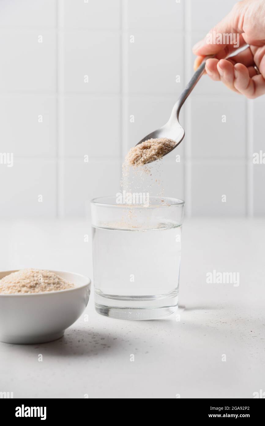La donna aggiunge fibra di psyllium al bicchiere d'acqua su uno sfondo bianco. Supercibo per intestini sani e dieta senza glutine. Foto Stock