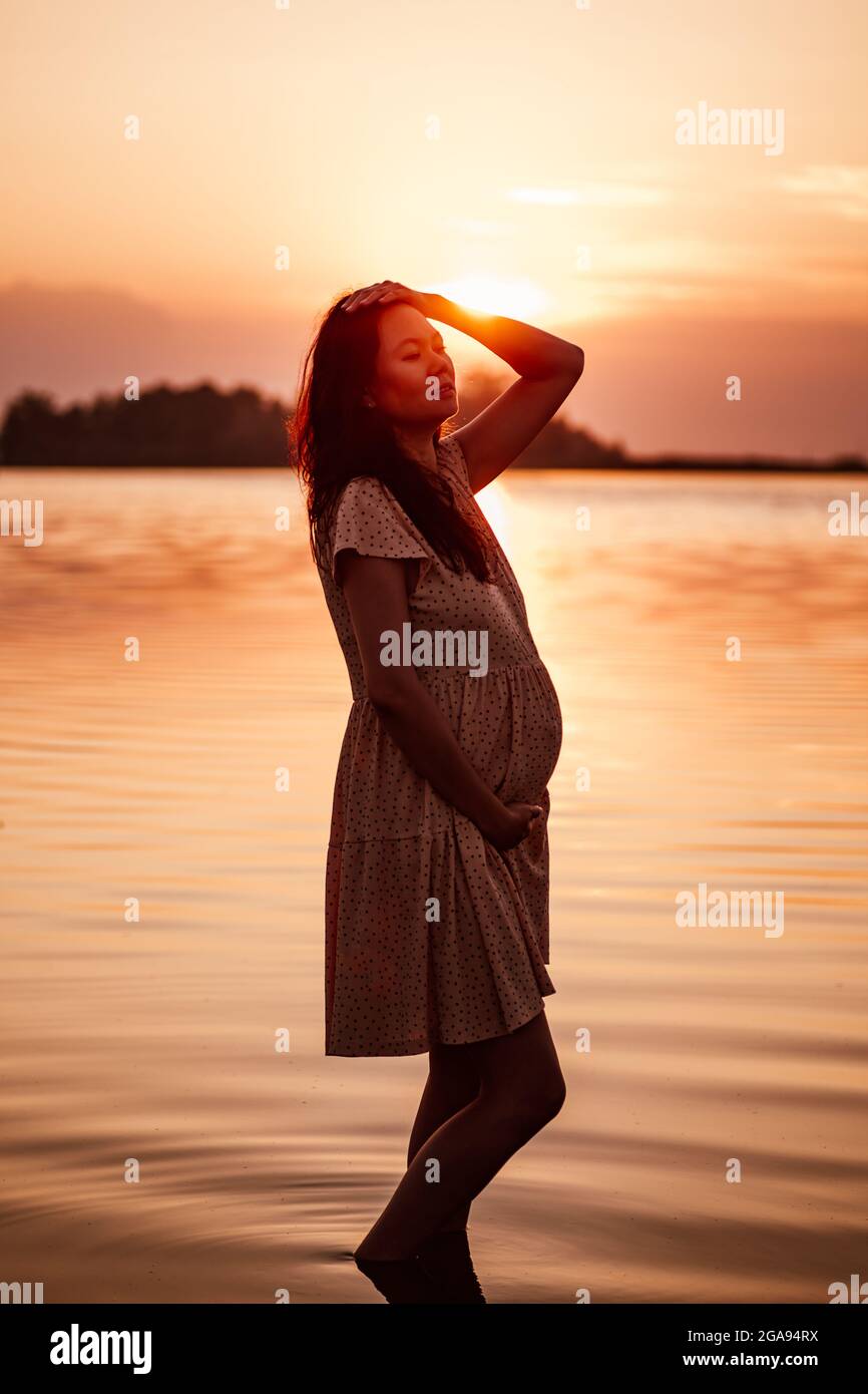 Donna incinta all'aperto. Silhouette di giovane donna felice in raggi di sole che si alza in piedi in acqua sulla riva del lago e che tocca i suoi capelli e stomaco Foto Stock