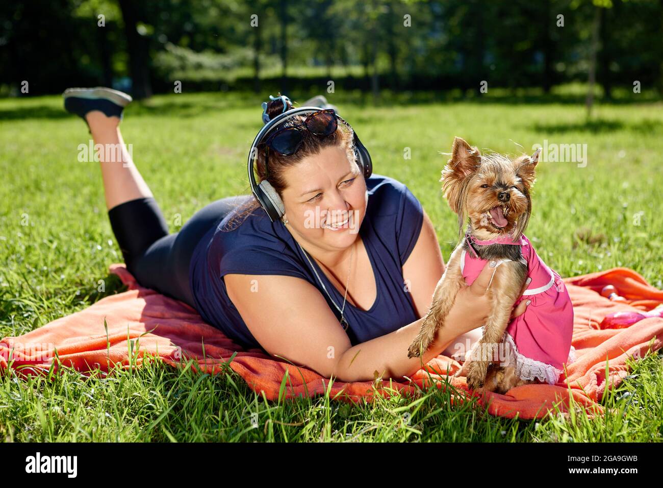 Donna in sovrappeso con cuffie wireless si trova nel parco con il cane e sorrisi. Foto Stock