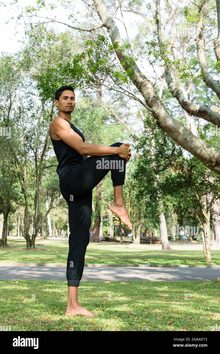 Giovane brasiliano che tira il ginocchio al petto, in un parco pubblico in una giornata di sole. Foto Stock