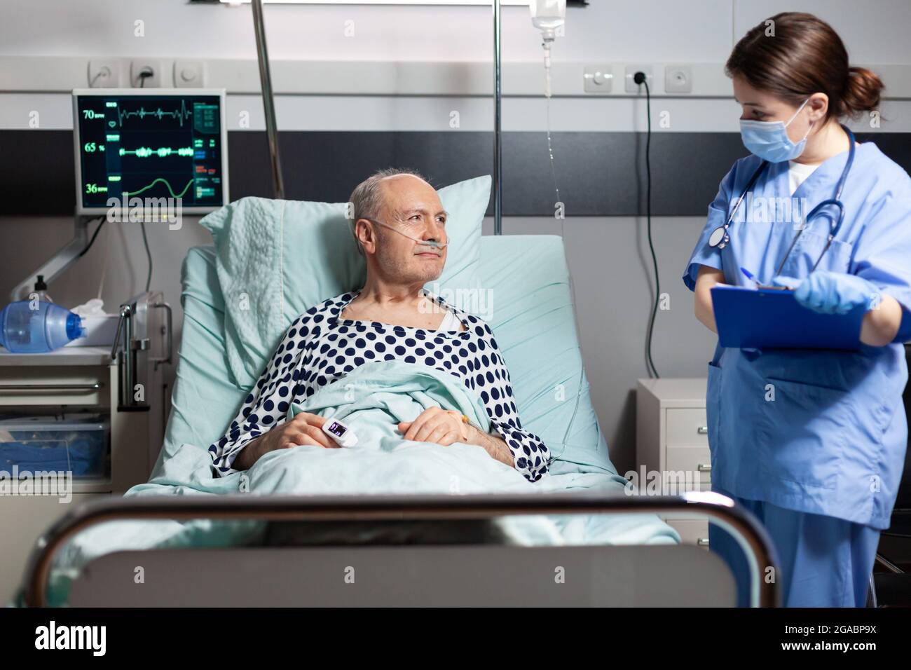 Infermiera medica in scrub con maschera chirurgica che prende notest sulla scacchiera durante la consultazione di malato uomo anziano malato. Paziente con respirazione fleboclisi ev con l'aiuto della maschera di ossigeno. Foto Stock