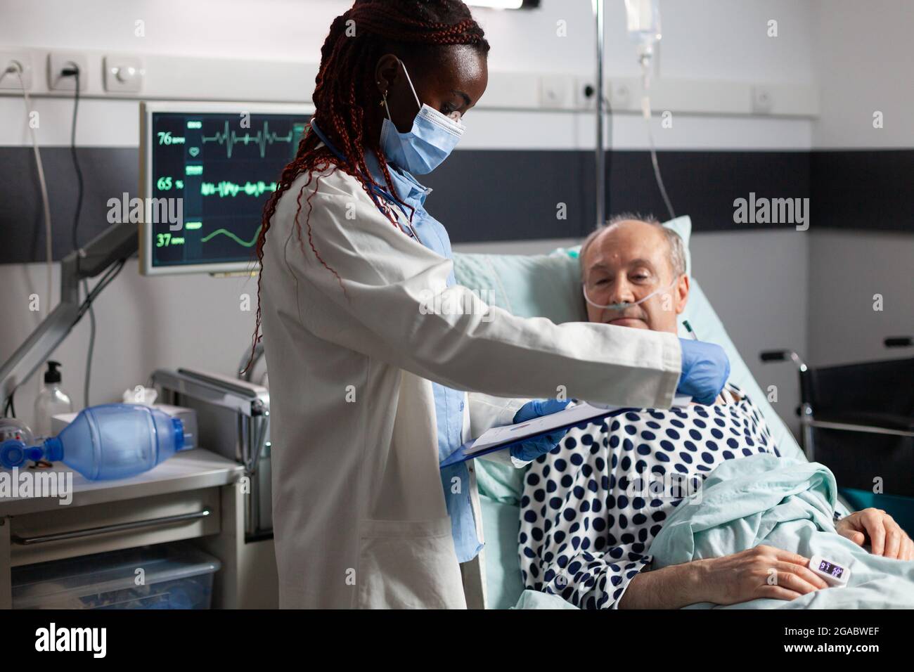 Medico afroamericano che indossa maschera chirurgica, in sala d'ospedale durante la consultazione di malato malato uomo anziano, respirando con l'aiuto di tubo di prova e fleboclisi iv attaccato. Foto Stock