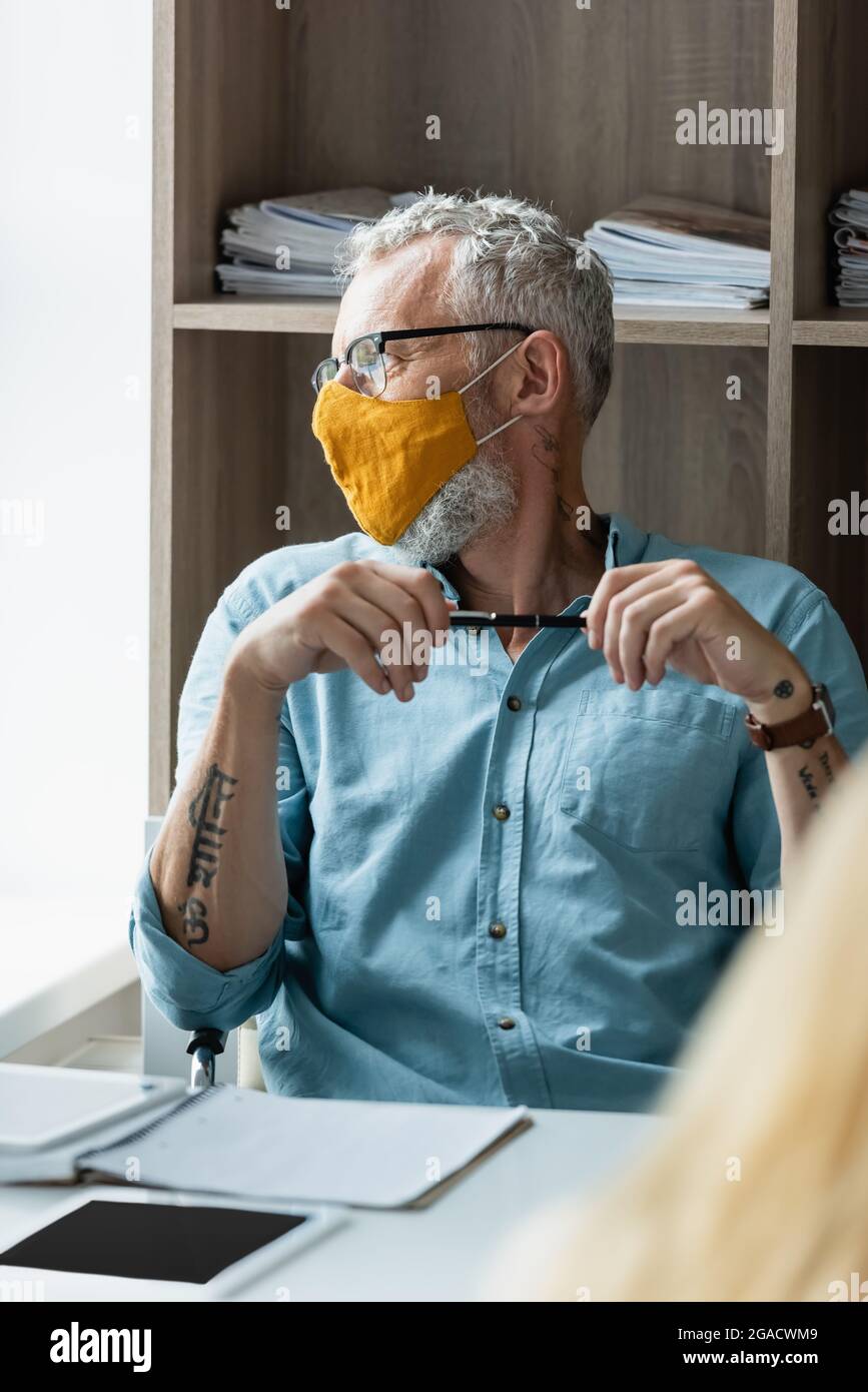 Insegnante in maschera protettiva che tiene la penna vicino a notebook e tablet digitali sul tavolo Foto Stock
