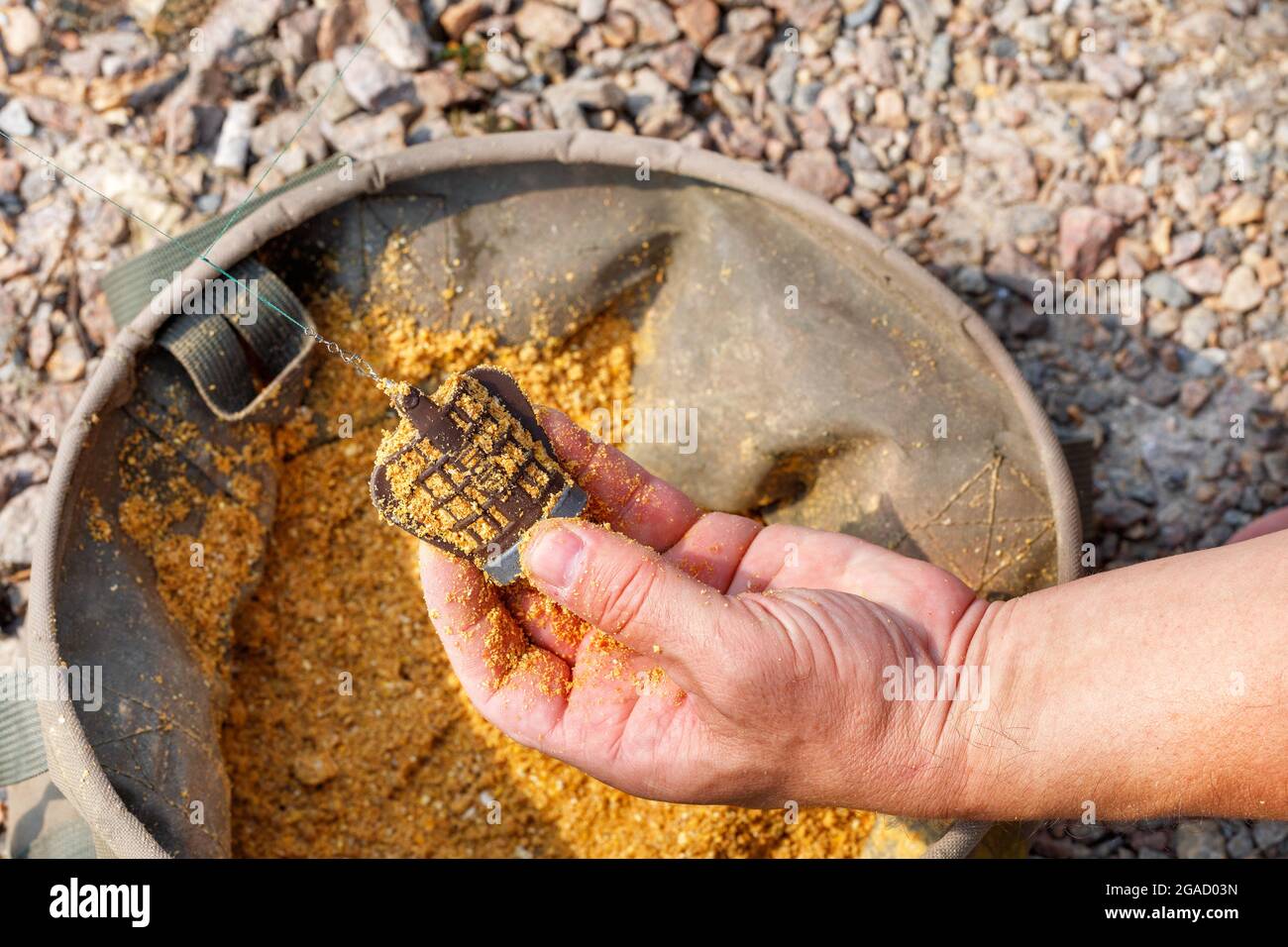 Nella mano di un pescatore un alimentatore con cibo di pesce sullo sfondo di un secchio di pesca tela in sfocatura e luce solare. Spazio di copia, primo piano. Foto Stock