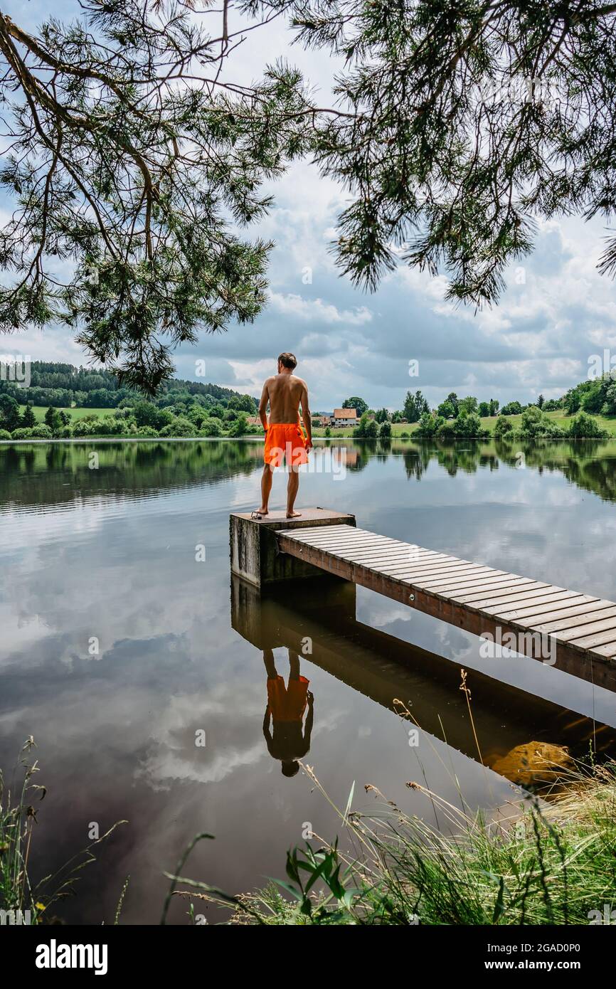 Vista posteriore di uomo che salta dal molo di legno con gioia in acqua, la natura riflessa in Lake.having estate divertimento da stagno.Travel vacanza Foto Stock
