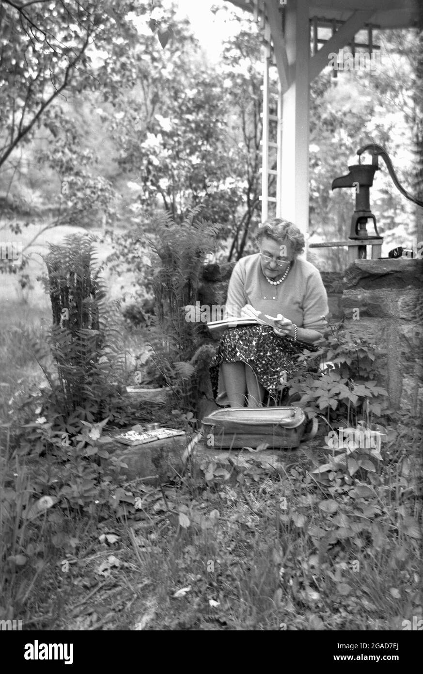 anni '50, storica, una signora anziana seduta fuori da un vecchio pozzo d'acqua pittura su un tampone di carta, forse facendo un acquerello. Foto Stock