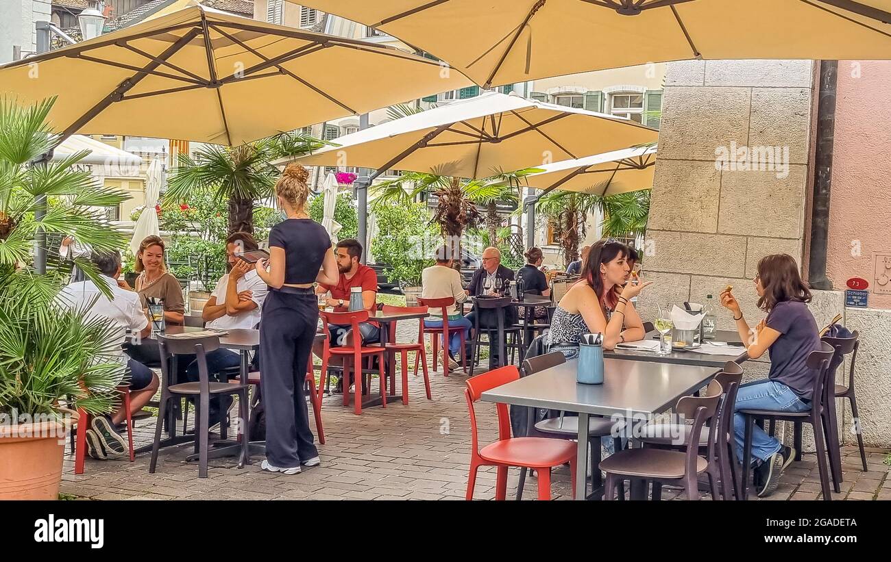 Gruppi seduti a tavola sotto ombrelloni all'aperto, in un cortile verde, in un bar di strada durante l'ora dell'aperitivo. Foto Stock