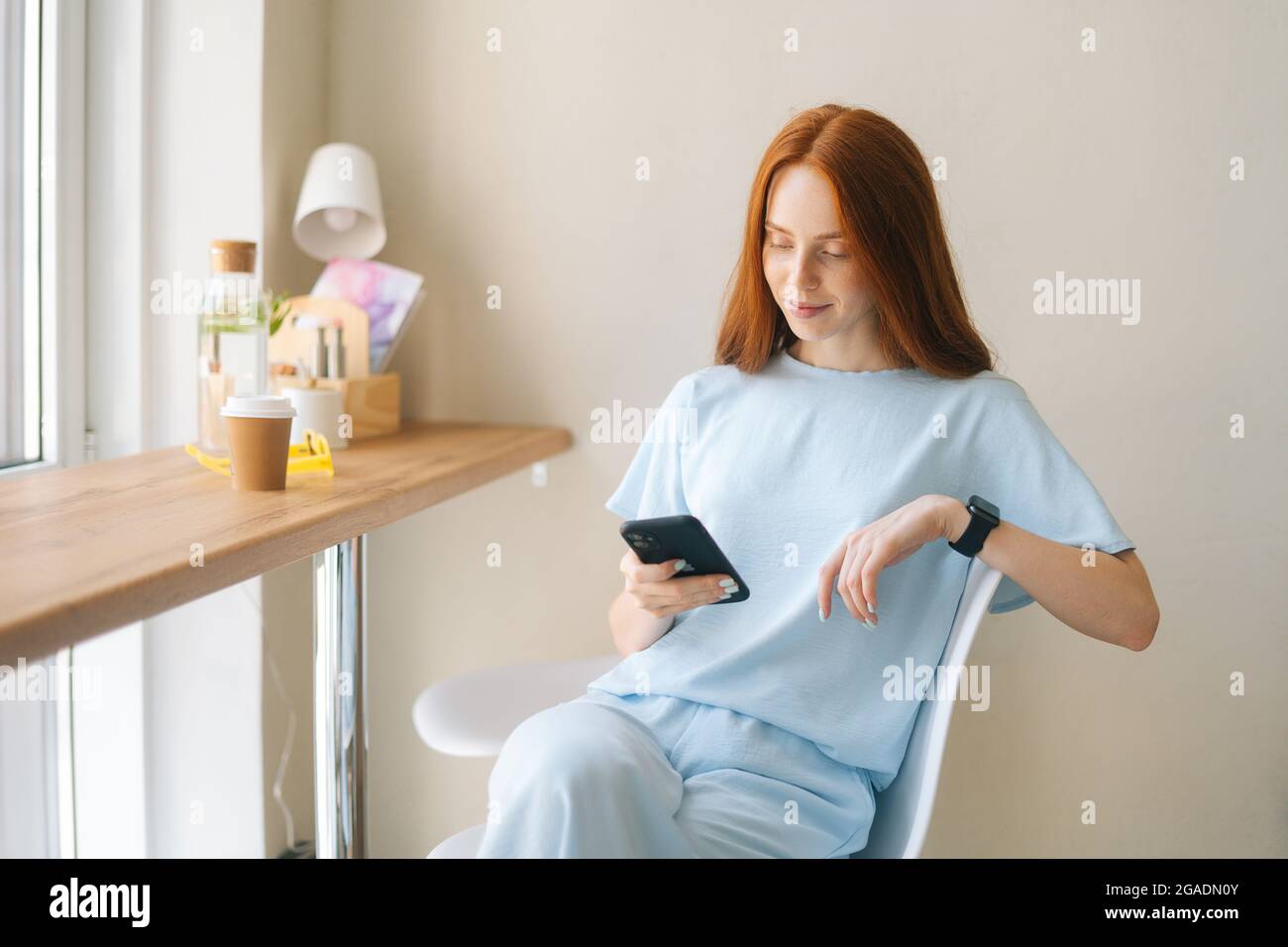 Foto media di adorabile giovane donna che indossa abiti casual utilizzando il telefono cellulare seduto alla scrivania accanto alla finestra in un accogliente e leggero caffè. Foto Stock